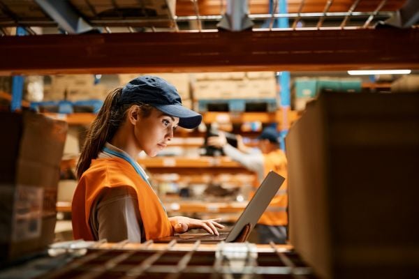 Image of a ecommerce fulfillment warehouse, looking past a shelf at a worker using a mobile computer to track inventory